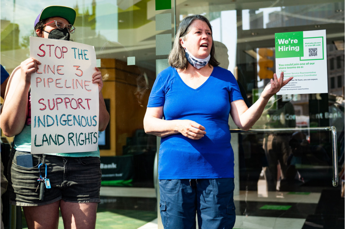 Person with glasses, hat and mask holds sign that says " Stop the Line 3 Pipeline, Support Inndigenous Rights" next to a white woman in a blue shirt, speaking.