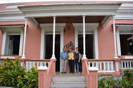 Three people stand in front of light pink house with porch