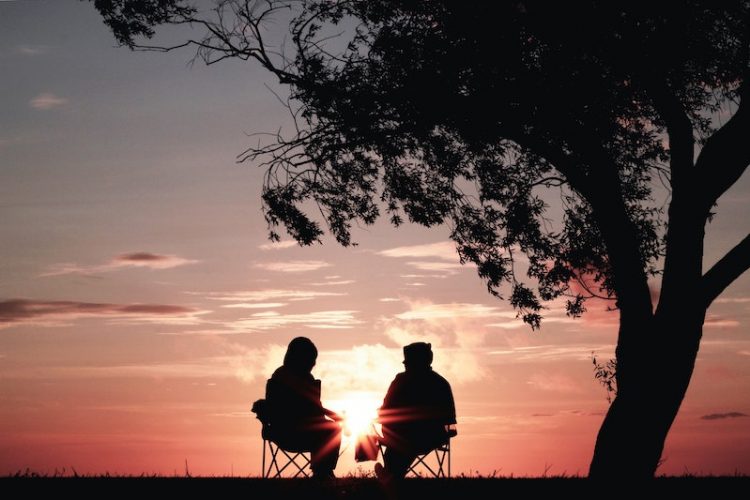 Two people backlit by pink sunset sitting in chairs under a tree