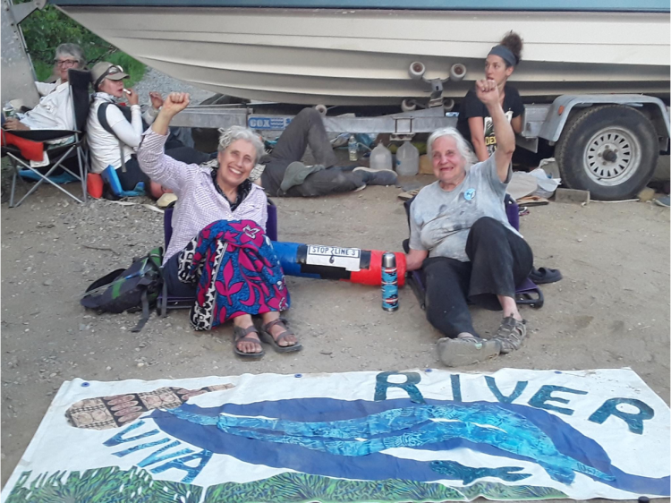 Two women with white hair raise hands in air while other arms are locked together in front of a sign that says "Viva River"