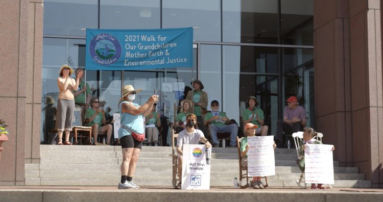 Twelve people sitting in rocking chairs or standing and clapping standing on steps in front of a bank. Sign in background says 2021 Walk for Our Grandchildren, Mother Earth, and Environmental Justice