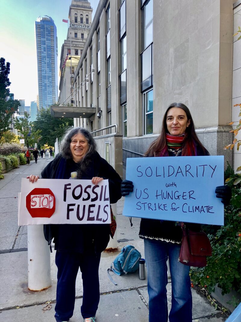 Two people standing in front of building with signs. One says "Stop Fossil Fuels." The other says, "Solidarity with the US Hunger Strike"