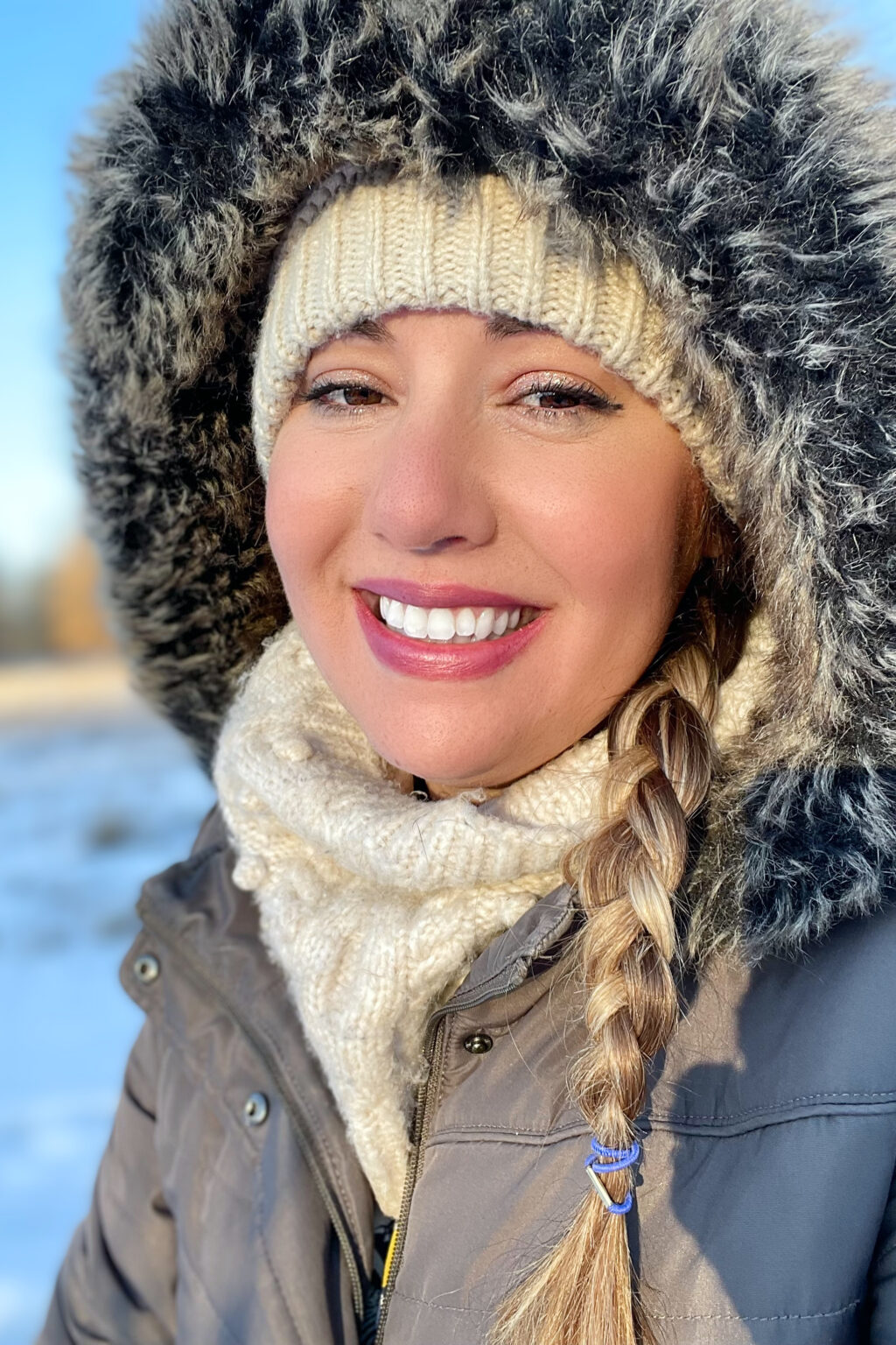 Person smiling with tan hat and scarf with grey jacket and snow in backgorund